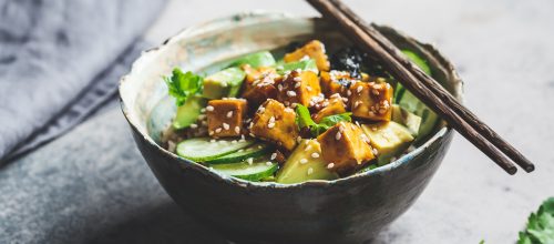Vegan tofu poke bowl with rice, cucumber, avocado and nori in a ceramic bowl, gray background.